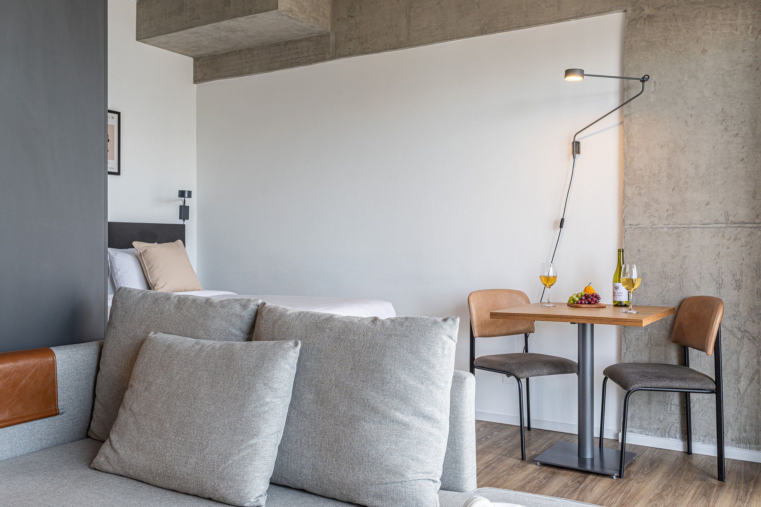 A modern hotel room with a sofa, two chairs, a small table with a bottle and glasses, and a lamp.