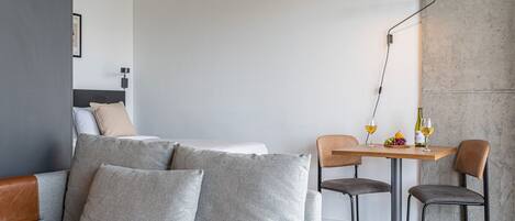 A modern hotel room with a sofa, two chairs, a small table with a bottle and glasses, and a lamp.