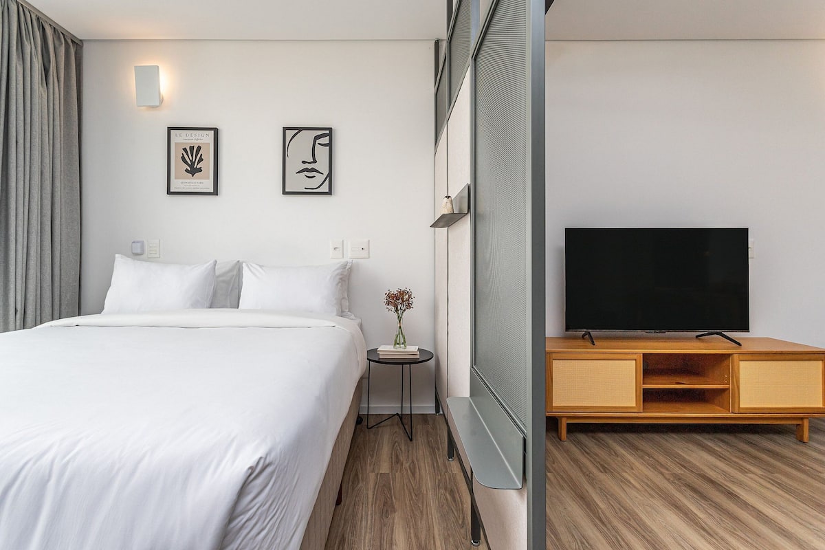 A hotel room with a bed, a small table with a vase, a TV on a wooden stand, and two framed artworks on the wall.