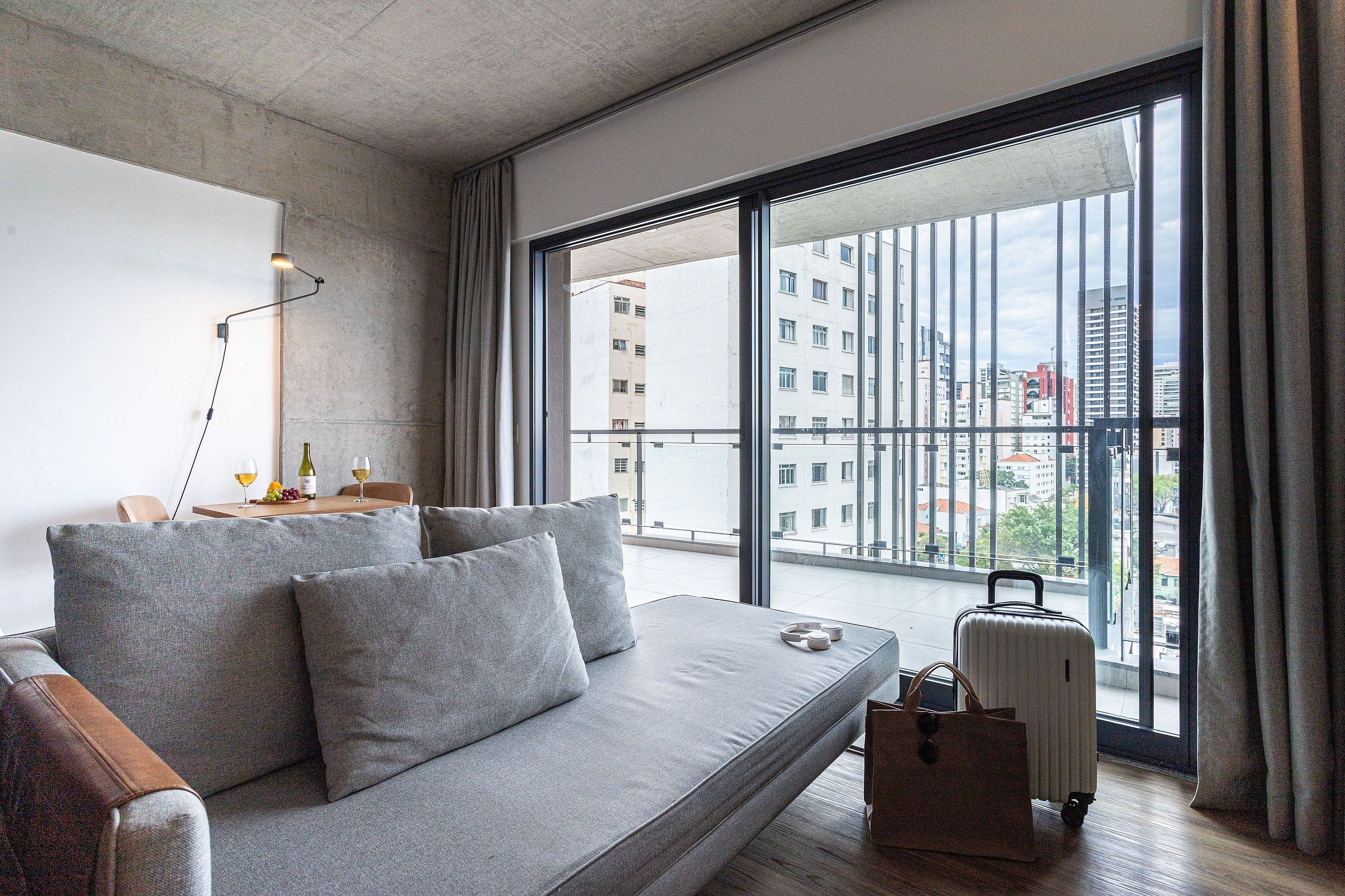 A modern living room with a grey sofa, a small table with a lamp, and a view of the city through large windows.