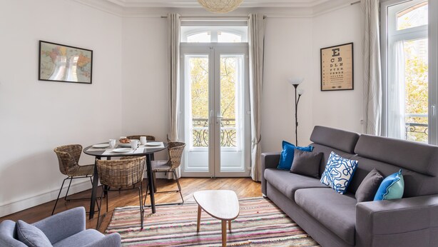 A living room with a grey sofa, a dining table with chairs, a large window with curtains, and a hanging light fixture. - Le Milady - Charmant appartement au cœur de Rennes (Rennes)