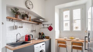 A compact kitchen with a sink, a washing machine, a red kettle, and a window with a view of buildings. - Le Milady - Charmant appartement au cœur de Rennes (Rennes)