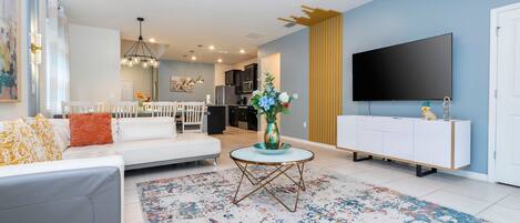 A modern living room with a grey sofa, white dining table, and a TV mounted on the wall.