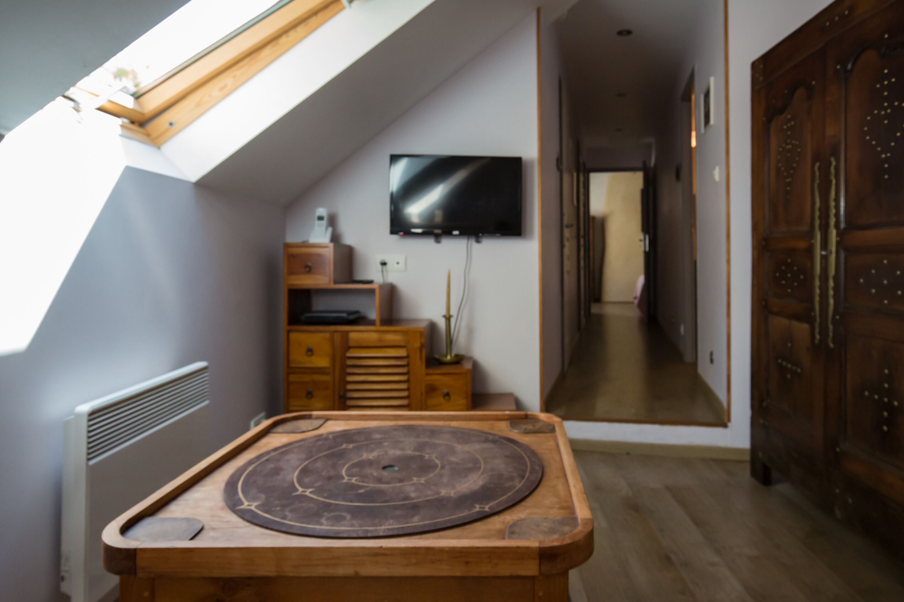 Een kamer met een schuine kap, een houten tafel, een aan de muur bevestigde televisie en een grote houten garderobe.