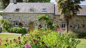 Ein Steinhaus mit Holztür, einem kleinen Fenster und einem Garten mit verschiedenen Pflanzen und Blumen.
