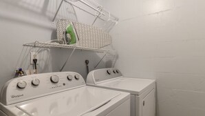 Uma lavanderia com duas máquinas de lavar e secar Whirlpool, uma prateleira de metal com um tecido estampado e uma tomada na parede.