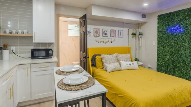 Quarto compacto com cama, uma pequena mesa com pratos, micro-ondas e parede verde com um letreiro neon.
