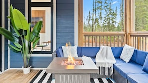 Un espace extérieur moderne, aménagé pour se détendre, avec un foyer, un pot de fleurs et une vue sur les arbres à travers une fenêtre.