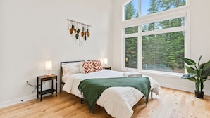 Une chambre avec une grande fenêtre, un lit avec une literie blanche et un plaid vert, une table de chevet avec une lampe, et une plante fixée au mur.
