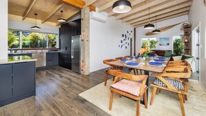Eine moderne Küche mit Holzbalken, einem Essbereich mit langem Tisch und Stühlen und Blick ins Freie durch große Fenster.