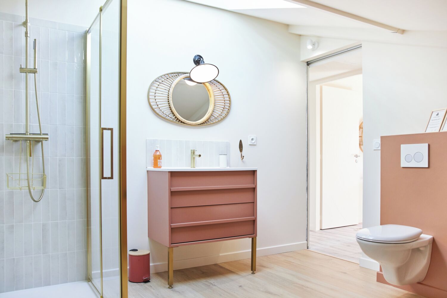 Een moderne badkamer met een douchecabine van glas, een roze wastafelmeubel met een ronde spiegel en een wit toilet.