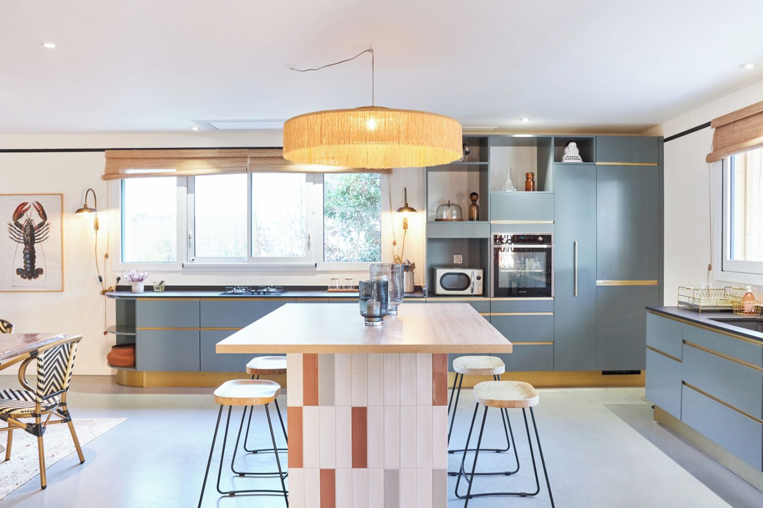 A modern kitchen with a central island, bar stools, a hanging light fixture, and a view of the outdoors through large windows.