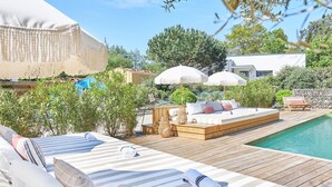 A wooden deck with a pool, a wooden bed with pillows, and two white umbrellas.