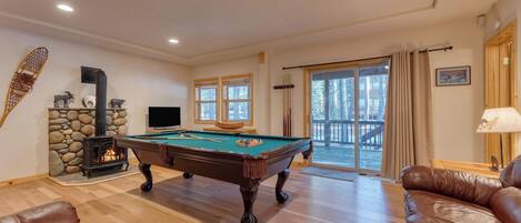 A cozy living room with a pool table, a fireplace, and a large window overlooking a deck.