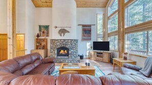 A cozy living room with a stone fireplace, leather sofas, a flat-screen TV, and large windows offering a forest view. - Hansel by Avantstay Pool Table, 5mins to Skiing (Truckee)