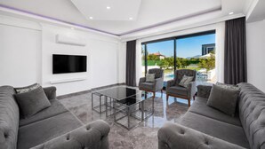 Ein modernes Wohnzimmer mit Flachbildfernseher, grauen Sofas, einem Glastisch und einem großen Fenster mit Blick auf einen Pool und Berge.