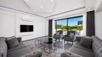 Ein modernes Wohnzimmer mit Flachbildfernseher, grauen Sofas, einem Glastisch und einem großen Fenster mit Blick auf einen Pool und Berge.