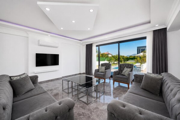 Ein modernes Wohnzimmer mit Flachbildfernseher, grauen Sofas, einem Glastisch und einem großen Fenster mit Blick auf einen Pool und Berge.