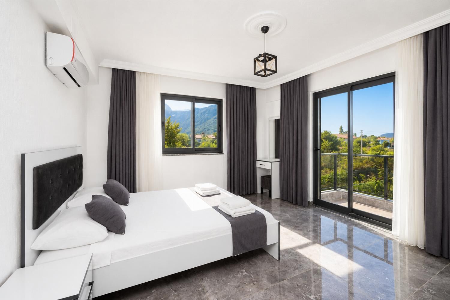 Une chambre d’hôtel moderne équipée d’un lit, d’un bureau, d’une chaise, d’une grande fenêtre et d’un balcon offrant une vue sur la verdure et les montagnes.