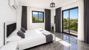 Ein modernes Hotelzimmer mit Bett, Schreibtisch, Sessel, großem Fenster und einem Balkon mit Blick auf Grünflächen und Berge.