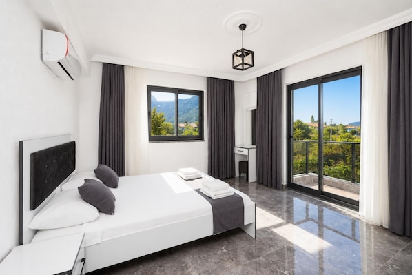 A modern hotel room with a bed, a desk, a chair, a large window, and a balcony with a view of greenery and mountains.