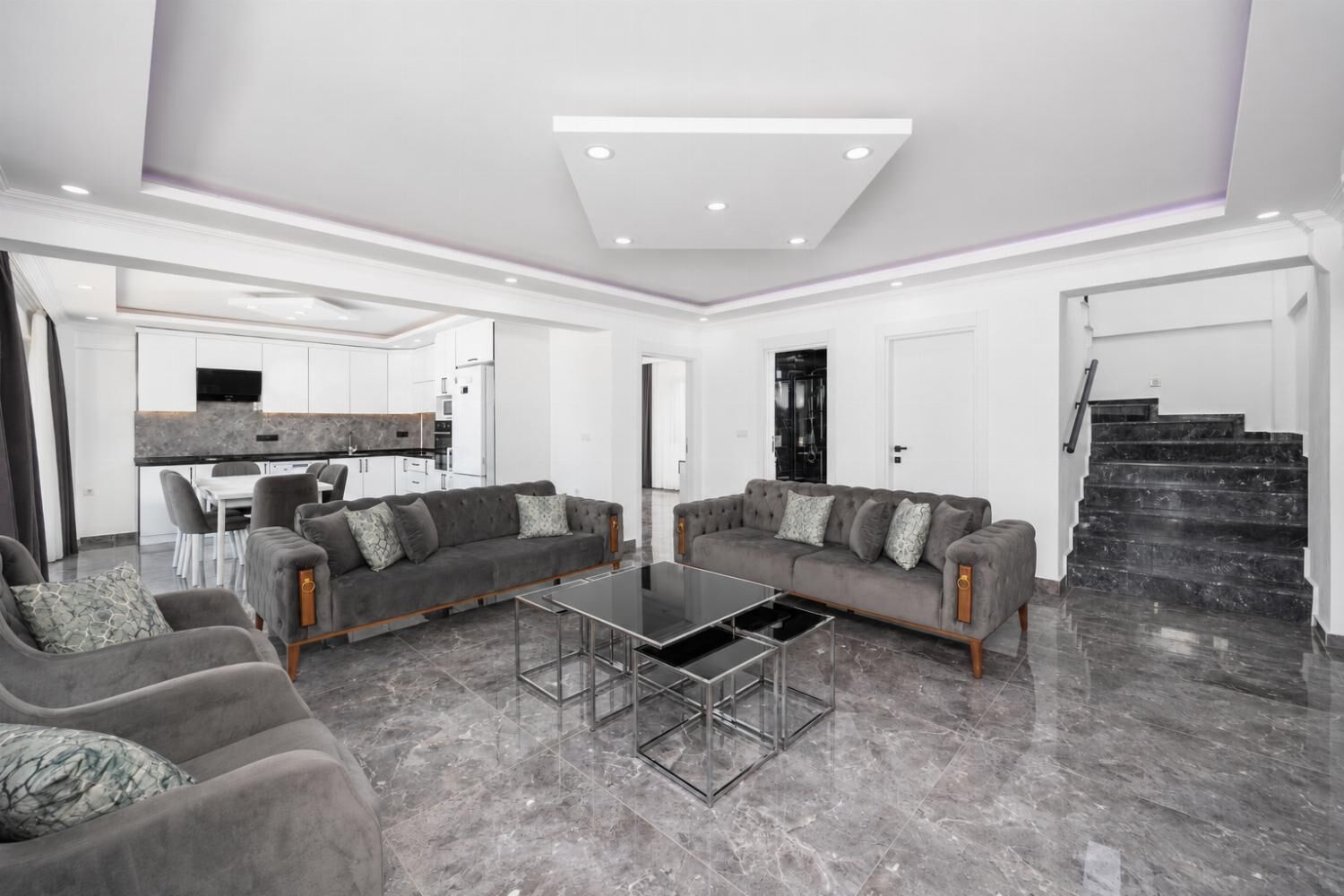 Un salon moderne avec deux canapés gris, une table basse en verre et une cuisine en arrière-plan.