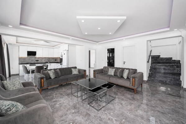 Ein modernes Wohnzimmer mit zwei grauen Sofas, einem Glastisch und einer Küche im Hintergrund.