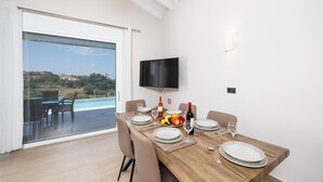 A dining area with a table set for four, a television mounted on the wall, and a view of a swimming pool and greenery outside. - Casa di Pietra - Two Bedroom Villa, Sleeps 4 (Lakithra)