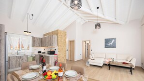 A modern kitchen with wooden cabinets, a dining area with a wooden table set for a meal, and a living area with a sofa and a painting on the wall. - Casa di Pietra - Two Bedroom Villa, Sleeps 4 (Lakithra)