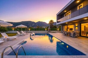 A swimming pool with lounge chairs and umbrellas, a patio area with outdoor furniture, and a mountain backdrop.