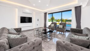 A modern living room with a flat-screen TV, two grey sofas, a glass coffee table, and a large window with a view of a swimming pool and mountains. - Villa Grand Yasar 1 - Four Bedroom Villa, Sleeps 8 (Okçular)