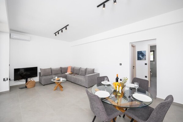 Een moderne woonkamer met een flatscreen televisie, een grijze bank en een glazen eettafel met stoelen.