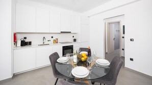 Een moderne keuken met witte kasten, een eethoek met een glazen tafel, en een klein kookeiland met fruit en een fles.