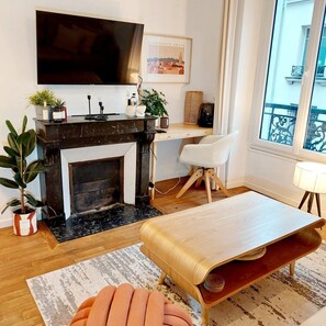 A modern living room with a fireplace, a flat-screen TV mounted above it, a desk with a chair, and a large window with a view of the city.