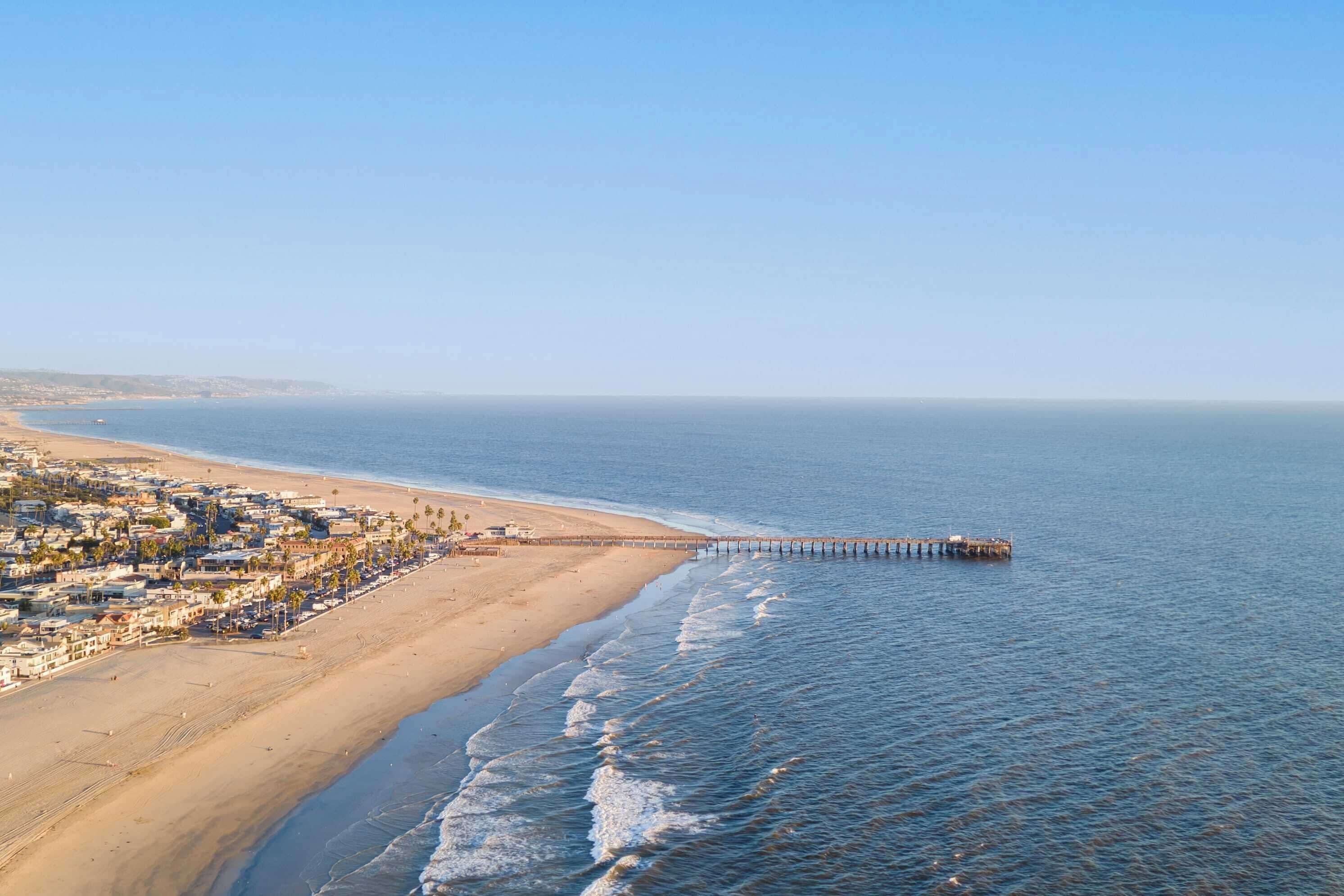 Escena costera con una playa, un muelle que se adentra en el agua, arena y una zona residencial con edificios.