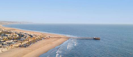 Escena costera con una playa, un muelle que se adentra en el agua, arena y una zona residencial con edificios.