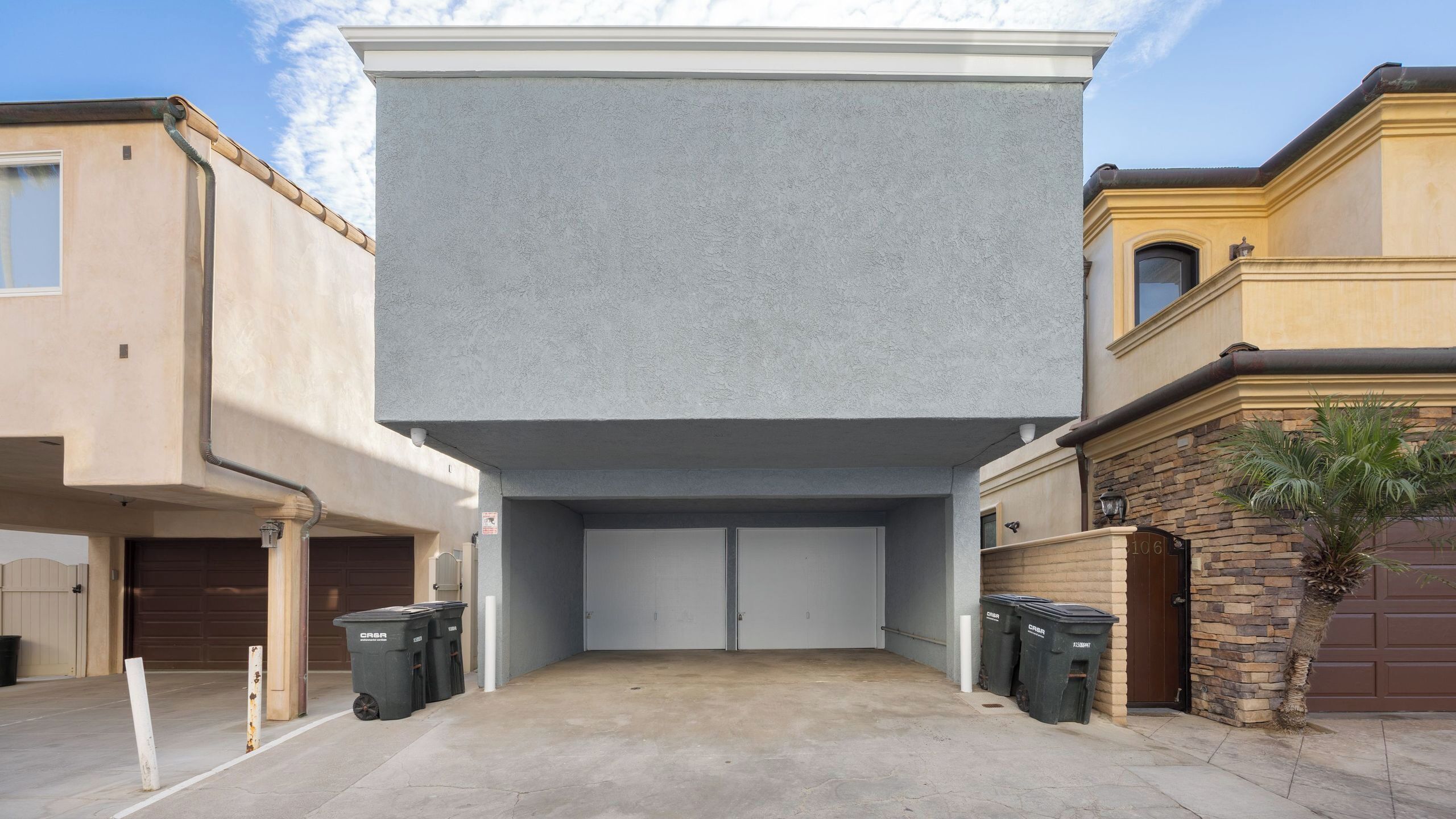 Un garage avec un grand mur à la texture marquée, deux poubelles et un palmier.
