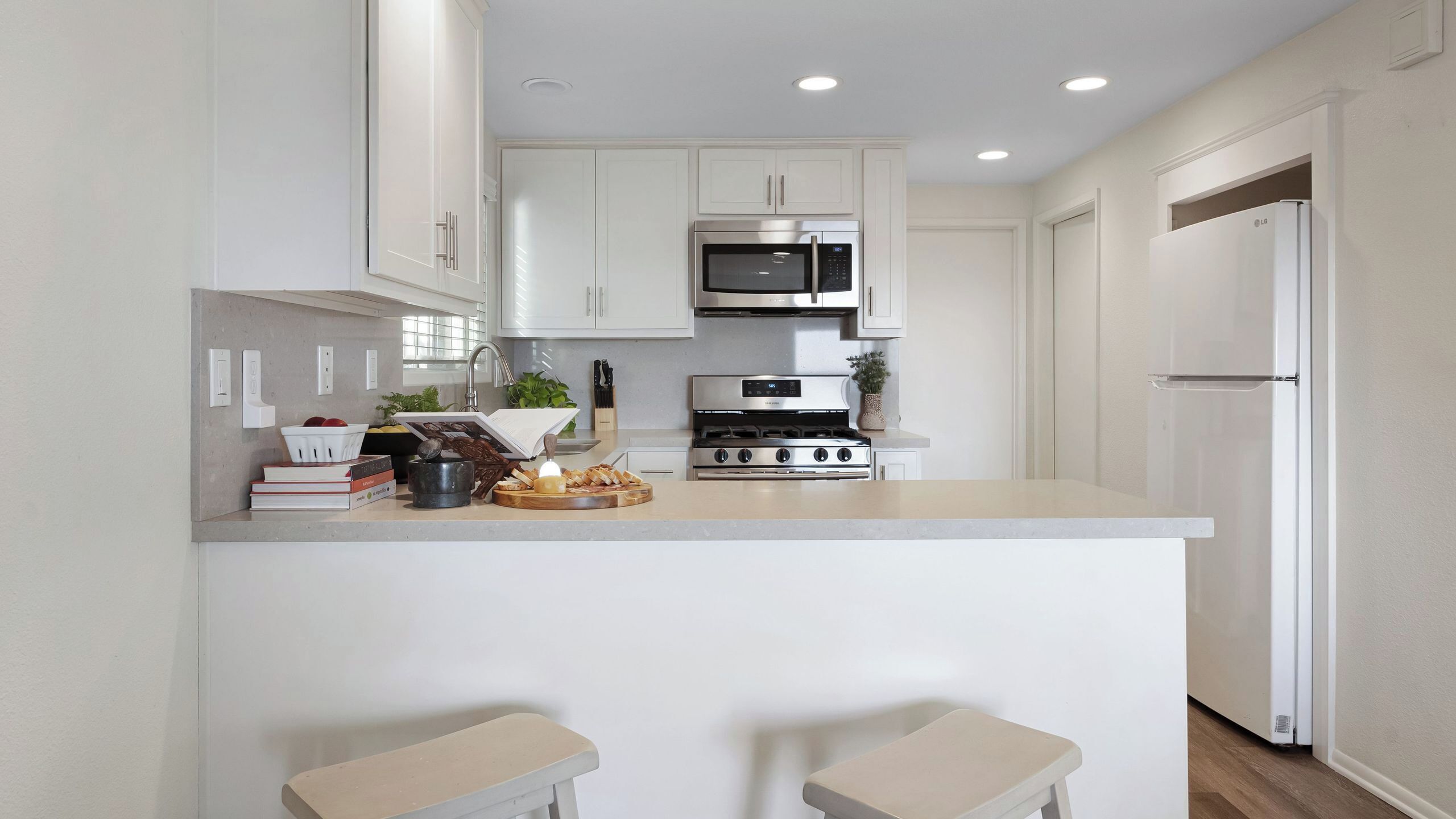 Una cocina moderna con armarios blancos, estufa de acero inoxidable, microondas y refrigerador. Hay dos taburetes en la encimera.