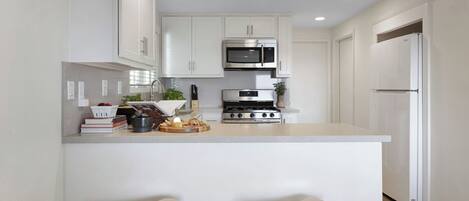 Una cocina moderna con armarios blancos, estufa de acero inoxidable, microondas y refrigerador. Hay dos taburetes en la encimera.