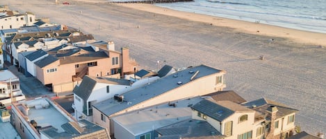 Un área residencial costera con casas a lo largo de la playa, un muelle al fondo y una vista despejada del océano.