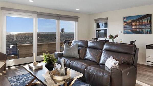 A modern living room with a leather sofa, a glass coffee table, and a view of the sea through large windows. - Balboa Breeze by Avantstay Beachfront Retreat + Ocean View (Newport Beach)