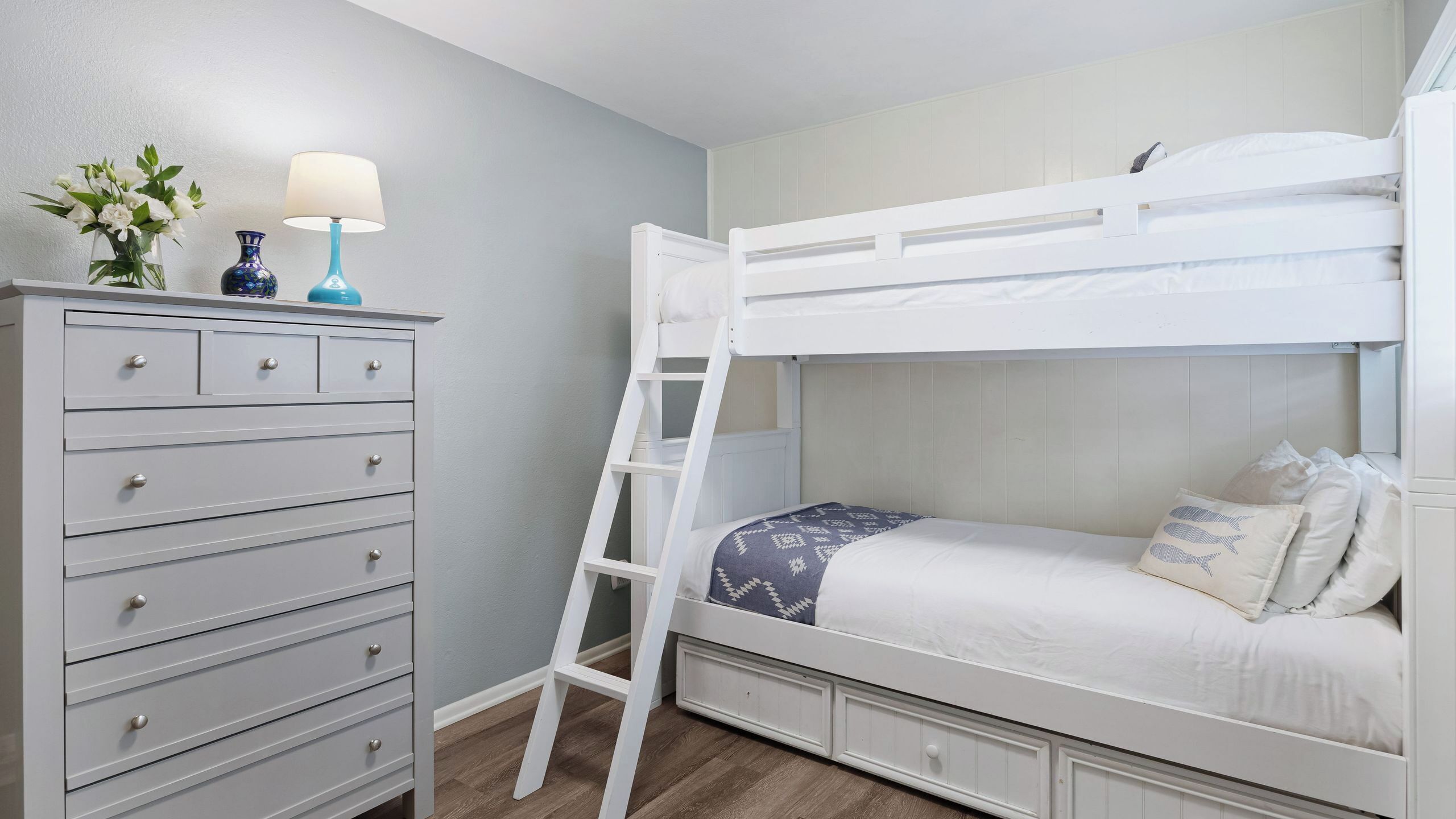 Una cama litera con escalera, un aparador con cajones y un florero con flores encima.