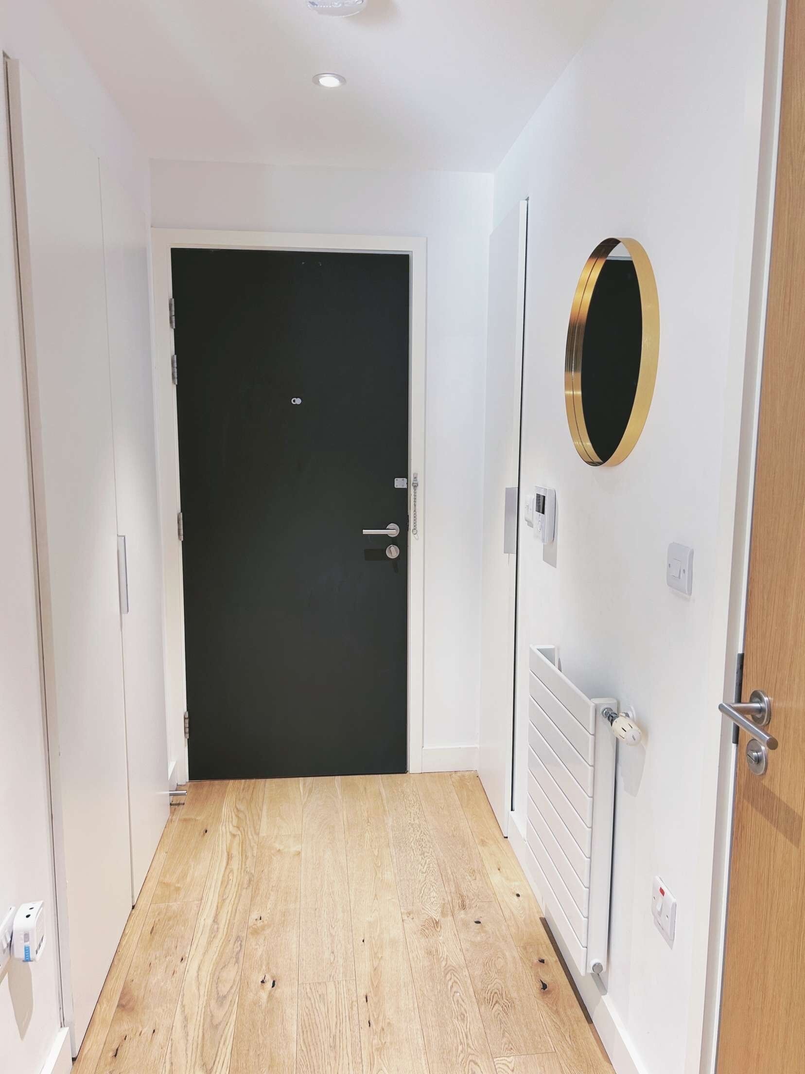 Un pasillo con piso de madera, una puerta negra y un espejo redondo en la pared.