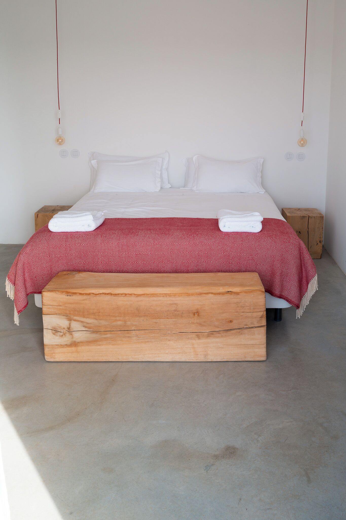 A bed with a wooden bench, white bedding, and a red patterned blanket.