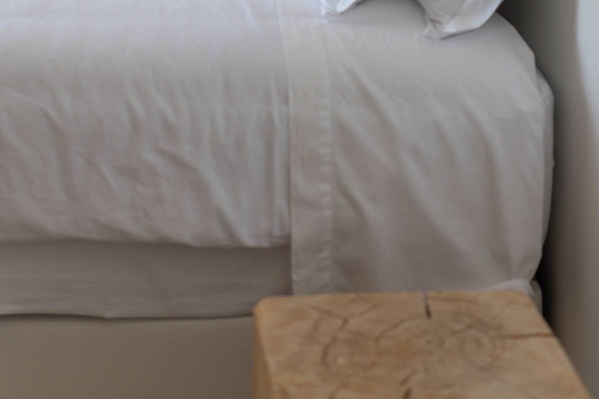A neatly made bed with white linens and pillows, a wooden bedside table, and a hanging light fixture.
