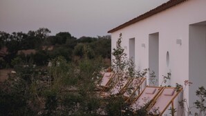 A white building with multiple rooms, each with a window and a door. There are wooden lounge chairs outside the building. - Alapega Live Slow (Alcacer do Sal)