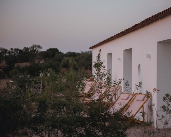 A white building with multiple rooms, each with a window and a door. There are wooden lounge chairs outside the building. - Alapega Live Slow (Alcacer do Sal)