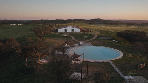 A large, circular swimming pool with a wooden deck and lounge chairs, surrounded by a white building and a landscaped garden. - Alapega Live Slow (Alcacer do Sal)