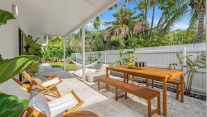 Une terrasse couverte avec une table et des chaises en bois, entourée de plantes tropicales et d’une clôture blanche.