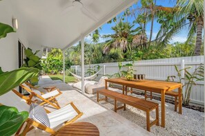 Eine überdachte Terrasse mit Holztisch und -stühlen, umgeben von tropischen Pflanzen und einem weißen Zaun.